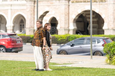 50 'li yaşlarda Malezyalı bir adam ve 30' lu yaşlarda Çinli bir kadın. İkisi de geleneksel Malezya kıyafeti giymiş. Malezya 'nın başkenti Kuala Lumpur' daki tarihi binanın önünde çimlerin üzerinde yan yana duruyorlar..