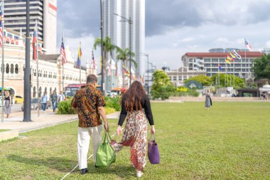 50 'li yaşlarda Malezyalı bir adam ve 30' lu yaşlarda Çinli bir kadın. İkisi de geleneksel Malezya kıyafeti giymiş. Malezya 'nın başkenti Kuala Lumpur' daki tarihi binanın önünde çimlerin üzerinde birlikte yürüyorlar..