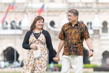 50 'li yaşlarda Malezyalı bir adam ve 30' lu yaşlarda Çinli bir kadın. İkisi de geleneksel Malezya kıyafeti giymiş. Malezya 'nın başkenti Kuala Lumpur' daki tarihi binanın önünde çimlerin üzerinde birlikte yürüyorlar..