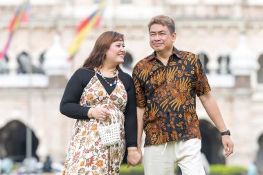 50 'li yaşlarda Malezyalı bir adam ve 30' lu yaşlarda Çinli bir kadın. İkisi de geleneksel Malezya kıyafeti giymiş. Malezya 'nın başkenti Kuala Lumpur' daki tarihi binanın önünde çimlerin üzerinde birlikte yürüyorlar..