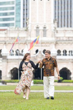 50 'li yaşlarda Malezyalı bir adam ve 30' lu yaşlarda Çinli bir kadın. İkisi de geleneksel Malezya kıyafeti giymiş. Malezya 'nın başkenti Kuala Lumpur' daki tarihi binanın önünde çimlerin üzerinde birlikte yürüyorlar..