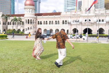 50 'li yaşlarda Malezyalı bir adam ve 30' lu yaşlarda Çinli bir kadın. İkisi de geleneksel Malezya kıyafeti giymiş. Malezya 'nın başkenti Kuala Lumpur' daki tarihi binanın önünde çimlerin üzerinde birlikte yürüyorlar..