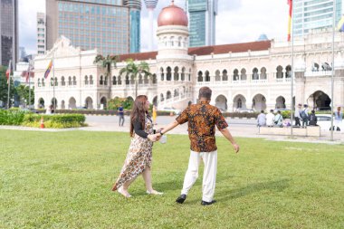 50 'li yaşlarda Malezyalı bir adam ve 30' lu yaşlarda Çinli bir kadın. İkisi de geleneksel Malezya kıyafeti giymiş. Malezya 'nın başkenti Kuala Lumpur' daki tarihi binanın önünde çimlerin üzerinde birlikte yürüyorlar..