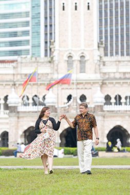 50 'li yaşlarda Malezyalı bir adam ve 30' lu yaşlarda Çinli bir kadın. İkisi de geleneksel Malezya kıyafeti giymiş. Malezya 'nın başkenti Kuala Lumpur' daki tarihi binanın önünde çimlerin üzerinde birlikte yürüyorlar..