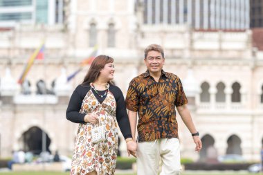 50 'li yaşlarda Malezyalı bir adam ve 30' lu yaşlarda Çinli bir kadın. İkisi de geleneksel Malezya kıyafeti giymiş. Malezya 'nın başkenti Kuala Lumpur' daki tarihi binanın önünde çimlerin üzerinde birlikte yürüyorlar..