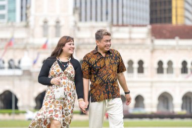 50 'li yaşlarda Malezyalı bir adam ve 30' lu yaşlarda Çinli bir kadın. İkisi de geleneksel Malezya kıyafeti giymiş. Malezya 'nın başkenti Kuala Lumpur' daki tarihi binanın önünde çimlerin üzerinde birlikte yürüyorlar..