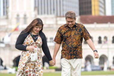 50 'li yaşlarda Malezyalı bir adam ve 30' lu yaşlarda Çinli bir kadın. İkisi de geleneksel Malezya kıyafeti giymiş. Malezya 'nın başkenti Kuala Lumpur' daki tarihi binanın önünde çimlerin üzerinde birlikte yürüyorlar..