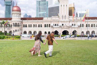 50 'li yaşlarda Malezyalı bir adam ve 30' lu yaşlarda Çinli bir kadın. İkisi de geleneksel Malezya kıyafeti giymiş. Malezya 'nın başkenti Kuala Lumpur' daki tarihi binanın önünde çimlerin üzerinde birlikte yürüyorlar..