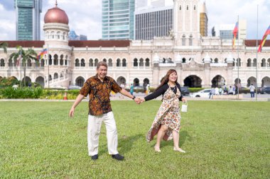 50 'li yaşlarda Malezyalı bir adam ve 30' lu yaşlarda Çinli bir kadın. İkisi de geleneksel Malezya kıyafeti giymiş. Malezya 'nın başkenti Kuala Lumpur' daki tarihi binanın önünde çimlerin üzerinde birlikte yürüyorlar..