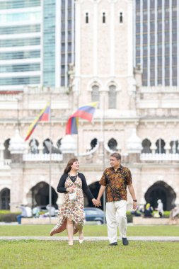 50 'li yaşlarda Malezyalı bir adam ve 30' lu yaşlarda Çinli bir kadın. İkisi de geleneksel Malezya kıyafeti giymiş. Malezya 'nın başkenti Kuala Lumpur' daki tarihi binanın önünde çimlerin üzerinde birlikte yürüyorlar..