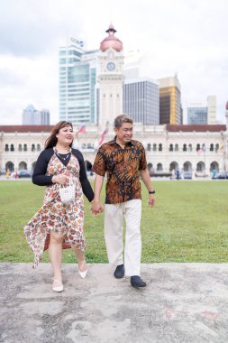 50 'li yaşlarda Malezyalı bir adam ve 30' lu yaşlarda Çinli bir kadın. İkisi de geleneksel Malezya kıyafeti giymiş. Malezya 'nın başkenti Kuala Lumpur' daki tarihi binanın önünde çimlerin üzerinde birlikte yürüyorlar..