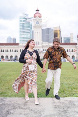 50 'li yaşlarda Malezyalı bir adam ve 30' lu yaşlarda Çinli bir kadın. İkisi de geleneksel Malezya kıyafeti giymiş. Malezya 'nın başkenti Kuala Lumpur' daki tarihi binanın önünde çimlerin üzerinde birlikte yürüyorlar..