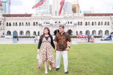 50 'li yaşlarda Malezyalı bir adam ve 30' lu yaşlarda Çinli bir kadın. İkisi de geleneksel Malezya kıyafeti giymiş. Malezya 'nın başkenti Kuala Lumpur' daki tarihi binanın önünde çimlerin üzerinde birlikte yürüyorlar..