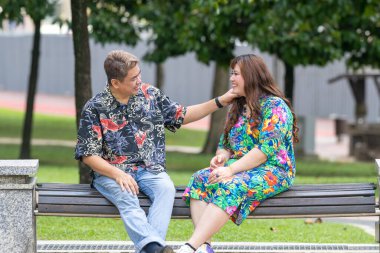 50 'li yaşlarda Malezyalı bir adam ve 30' lu yaşlarında Çinli bir kadın, ikisi de renkli giyinmiş, yakın bir bankta oturuyor ve Malezya 'nın Kuala Lumpur kentindeki KLCC Parkı' nda birlikte yürüyorlar..