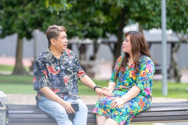 50 'li yaşlarda Malezyalı bir adam ve 30' lu yaşlarında Çinli bir kadın, ikisi de renkli giyinmiş, yakın bir bankta oturuyor ve Malezya 'nın Kuala Lumpur kentindeki KLCC Parkı' nda birlikte yürüyorlar..