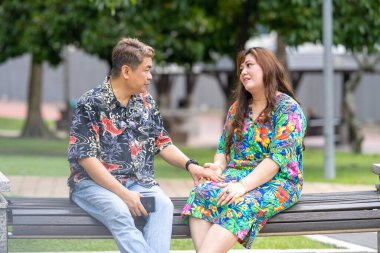 50 'li yaşlarda Malezyalı bir adam ve 30' lu yaşlarında Çinli bir kadın, ikisi de renkli giyinmiş, yakın bir bankta oturuyor ve Malezya 'nın Kuala Lumpur kentindeki KLCC Parkı' nda birlikte yürüyorlar..