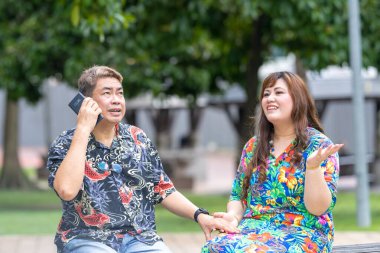 50 'li yaşlarda Malezyalı bir adam ve 30' lu yaşlarında Çinli bir kadın, ikisi de renkli giyinmiş, yakın bir bankta oturuyor ve Malezya 'nın Kuala Lumpur kentindeki KLCC Parkı' nda birlikte yürüyorlar..