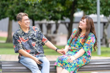 50 'li yaşlarda Malezyalı bir adam ve 30' lu yaşlarında Çinli bir kadın, ikisi de renkli giyinmiş, yakın bir bankta oturuyor ve Malezya 'nın Kuala Lumpur kentindeki KLCC Parkı' nda birlikte yürüyorlar..