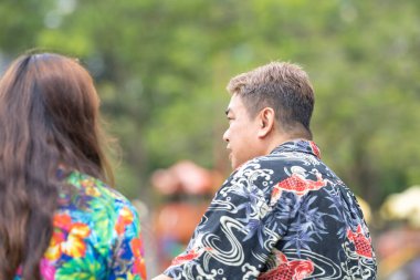 50 'li yaşlarda Malezyalı bir adam ve 30' lu yaşlarında Çinli bir kadın, ikisi de renkli giyinmiş, yakın bir bankta oturuyor ve Malezya 'nın Kuala Lumpur kentindeki KLCC Parkı' nda birlikte yürüyorlar..