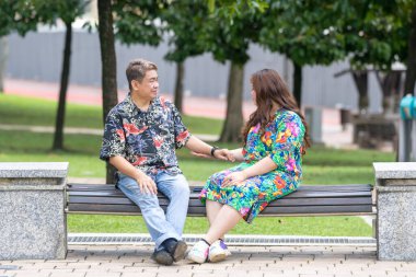 50 'li yaşlarda Malezyalı bir adam ve 30' lu yaşlarında Çinli bir kadın, ikisi de renkli giyinmiş, yakın bir bankta oturuyor ve Malezya 'nın Kuala Lumpur kentindeki KLCC Parkı' nda birlikte yürüyorlar..