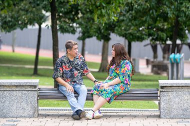 50 'li yaşlarda Malezyalı bir adam ve 30' lu yaşlarında Çinli bir kadın, ikisi de renkli giyinmiş, yakın bir bankta oturuyor ve Malezya 'nın Kuala Lumpur kentindeki KLCC Parkı' nda birlikte yürüyorlar..