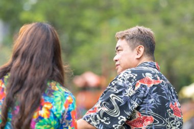 50 'li yaşlarda Malezyalı bir adam ve 30' lu yaşlarında Çinli bir kadın, ikisi de renkli giyinmiş, yakın bir bankta oturuyor ve Malezya 'nın Kuala Lumpur kentindeki KLCC Parkı' nda birlikte yürüyorlar..