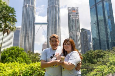 50 'li yaşlardaki Malezyalı adam açık mavi tişört giyiyor ve büyük Mickey Mouse baskısı ve 30' lu yaşlarında Çinli kadın KLCC Park 'ta gökdelenler Kuala Lumpur, Malezya.