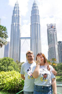 50 'li yaşlardaki Malezyalı adam açık mavi tişört giyiyor ve büyük Mickey Mouse baskısı ve 30' lu yaşlarında Çinli kadın KLCC Park 'ta gökdelenler Kuala Lumpur, Malezya.