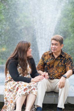 50 'li yaşlarda Malezyalı bir adam geleneksel 30' lu yaşlarda geleneksel bir elbise giyen Çinli bir kadın Kuala Lumpur 'daki Botani Perdana çeşmesinin önünde yan yana oturuyor.