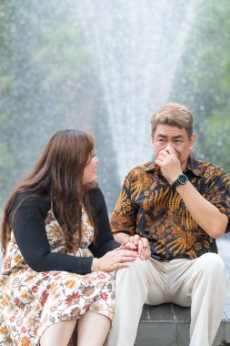 50 'li yaşlarda Malezyalı bir adam geleneksel 30' lu yaşlarda geleneksel bir elbise giyen Çinli bir kadın Kuala Lumpur 'daki Botani Perdana çeşmesinin önünde yan yana oturuyor.