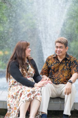 50 'li yaşlarda Malezyalı bir adam geleneksel 30' lu yaşlarda geleneksel bir elbise giyen Çinli bir kadın Kuala Lumpur 'daki Botani Perdana çeşmesinin önünde yan yana oturuyor.