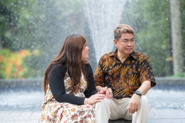 50 'li yaşlarda Malezyalı bir adam geleneksel 30' lu yaşlarda geleneksel bir elbise giyen Çinli bir kadın Kuala Lumpur 'daki Botani Perdana çeşmesinin önünde yan yana oturuyor.