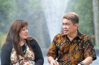 50 'li yaşlarda Malezyalı bir adam geleneksel 30' lu yaşlarda geleneksel bir elbise giyen Çinli bir kadın Kuala Lumpur 'daki Botani Perdana çeşmesinin önünde yan yana oturuyor.