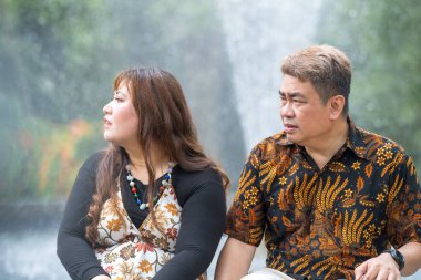 50 'li yaşlarda Malezyalı bir adam geleneksel 30' lu yaşlarda geleneksel bir elbise giyen Çinli bir kadın Kuala Lumpur 'daki Botani Perdana çeşmesinin önünde yan yana oturuyor.