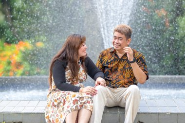 50 'li yaşlarda Malezyalı bir adam geleneksel 30' lu yaşlarda geleneksel bir elbise giyen Çinli bir kadın Kuala Lumpur 'daki Botani Perdana çeşmesinin önünde yan yana oturuyor.