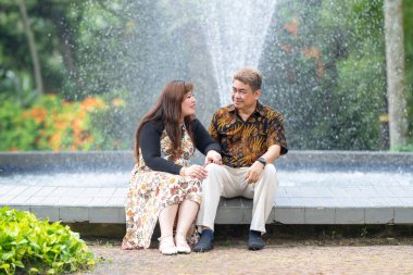 50 'li yaşlarda Malezyalı bir adam geleneksel 30' lu yaşlarda geleneksel bir elbise giyen Çinli bir kadın Kuala Lumpur 'daki Botani Perdana çeşmesinin önünde yan yana oturuyor.