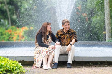 50 'li yaşlarda Malezyalı bir adam geleneksel 30' lu yaşlarda geleneksel bir elbise giyen Çinli bir kadın Kuala Lumpur 'daki Botani Perdana çeşmesinin önünde yan yana oturuyor.