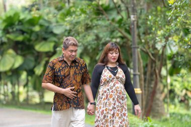 50 'li yaşlarda Malezyalı bir adam geleneksel Malezya gömleği giyiyor ve 30' lu yaşlarda Çinli bir kadın geleneksel elbiseler giyiyor, Taman Botani Perdana, Kuala Lumpur, Malezya 'da bitkilerle çevrili yürüyor..