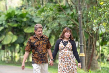 50 'li yaşlarda Malezyalı bir adam geleneksel Malezya gömleği giyiyor ve 30' lu yaşlarda Çinli bir kadın geleneksel elbiseler giyiyor, Taman Botani Perdana, Kuala Lumpur, Malezya 'da bitkilerle çevrili yürüyor..