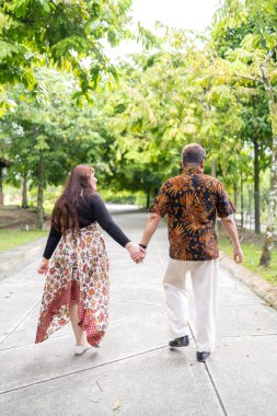 50 'li yaşlarda Malezyalı bir adam geleneksel Malezya gömleği giyiyor ve 30' lu yaşlarda Çinli bir kadın geleneksel elbiseler giyiyor, Taman Botani Perdana, Kuala Lumpur, Malezya 'da bitkilerle çevrili yürüyor..