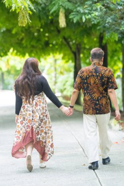 50 'li yaşlarda Malezyalı bir adam geleneksel Malezya gömleği giyiyor ve 30' lu yaşlarda Çinli bir kadın geleneksel elbiseler giyiyor, Taman Botani Perdana, Kuala Lumpur, Malezya 'da bitkilerle çevrili yürüyor..