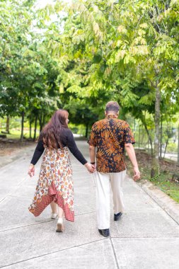 50 'li yaşlarda Malezyalı bir adam geleneksel Malezya gömleği giyiyor ve 30' lu yaşlarda Çinli bir kadın geleneksel elbiseler giyiyor, Taman Botani Perdana, Kuala Lumpur, Malezya 'da bitkilerle çevrili yürüyor..