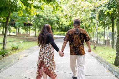 50 'li yaşlarda Malezyalı bir adam geleneksel Malezya gömleği giyiyor ve 30' lu yaşlarda Çinli bir kadın geleneksel elbiseler giyiyor, Taman Botani Perdana, Kuala Lumpur, Malezya 'da bitkilerle çevrili yürüyor..
