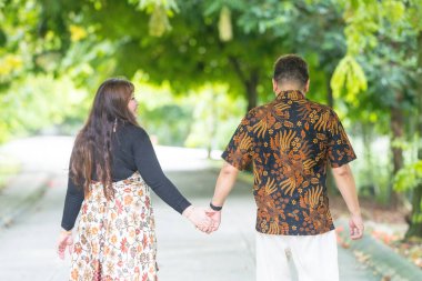 50 'li yaşlarda Malezyalı bir adam geleneksel Malezya gömleği giyiyor ve 30' lu yaşlarda Çinli bir kadın geleneksel elbiseler giyiyor, Taman Botani Perdana, Kuala Lumpur, Malezya 'da bitkilerle çevrili yürüyor..