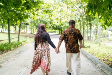 50 'li yaşlarda Malezyalı bir adam geleneksel Malezya gömleği giyiyor ve 30' lu yaşlarda Çinli bir kadın geleneksel elbiseler giyiyor, Taman Botani Perdana, Kuala Lumpur, Malezya 'da bitkilerle çevrili yürüyor..