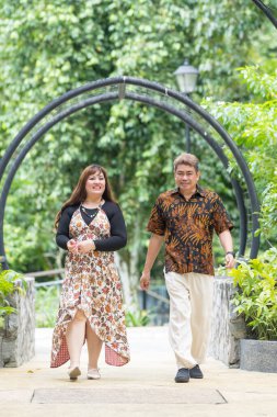 50 'li yaşlarda Malezyalı bir adam geleneksel Malezya gömleği giyiyor ve 30' lu yaşlarda Çinli bir kadın geleneksel elbiseler giyiyor, Taman Botani Perdana, Kuala Lumpur, Malezya 'da bitkilerle çevrili yürüyor..