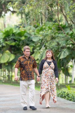 50 'li yaşlarda Malezyalı bir adam geleneksel Malezya gömleği giyiyor ve 30' lu yaşlarda Çinli bir kadın geleneksel elbiseler giyiyor, Taman Botani Perdana, Kuala Lumpur, Malezya 'da bitkilerle çevrili yürüyor..