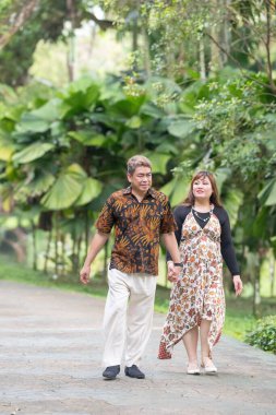 50 'li yaşlarda Malezyalı bir adam geleneksel Malezya gömleği giyiyor ve 30' lu yaşlarda Çinli bir kadın geleneksel elbiseler giyiyor, Taman Botani Perdana, Kuala Lumpur, Malezya 'da bitkilerle çevrili yürüyor..