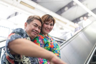 50 'li yaşlarında Malezyalı bir adam renkli gömlek giyiyor ve 30' lu yaşlarında Çinli bir kadın renkli elbiseler giyiyor, Malezya, Kuala Lumpur 'da bir tren istasyonunda birlikte vakit geçiriyorlar..