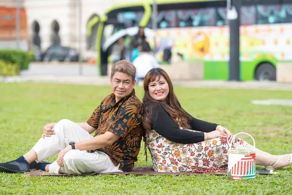 50 'li yaşlarda Malezyalı bir adam ve 30' lu yaşlarda Çinli bir kadın. İkisi de Malezya 'nın başkenti Kuala Lumpur' da bulunan tarihi bir binanın önündeki çimlerin üzerinde oturuyorlar..